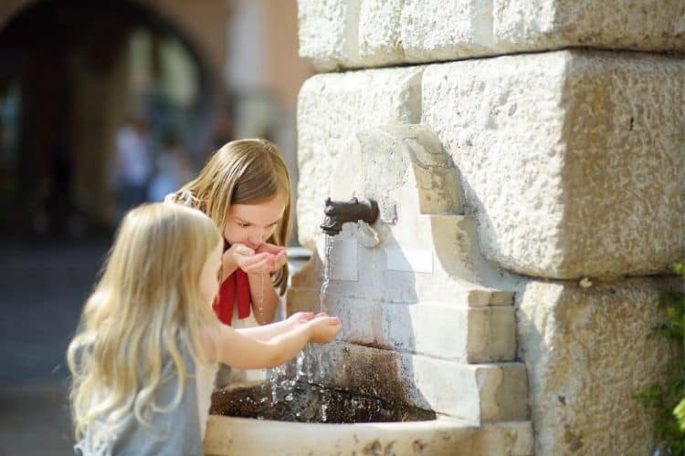 Two Cute Little Girls Playing with a Drinking Water Fountain on Warm and Sunny Summer Day